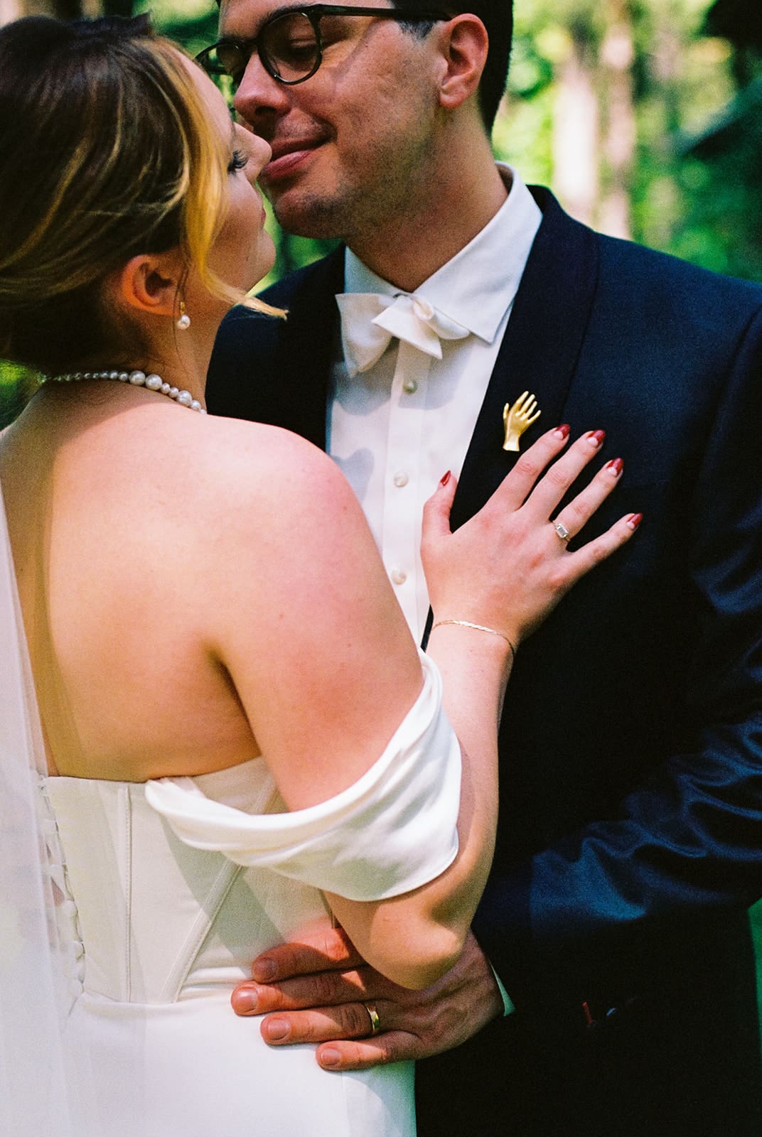 Close-up of a bride and groom in an intimate moment on their wedding day. The bride wears an off-the-shoulder white dress with a pearl necklace and red nails, gently resting her hand on the groom’s chest. The groom wears a dark suit with a white bow tie and a gold hand-shaped pin. They are smiling and leaning in, surrounded by soft natural light in a forested setting